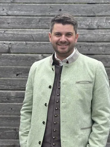 Philipp in front of a wooden wall, smiling, in a green jacket